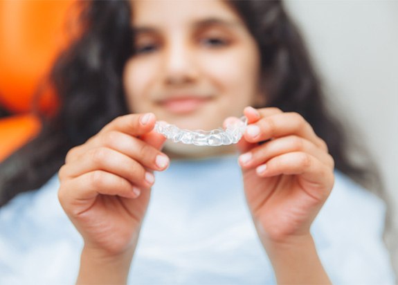 Teen girl smiling while holding clear aligner in treatment chair